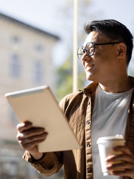 Asian businessman drinking coffee and watching something on digital tablet inside unknown building. Concept of remote and freelance work. Modern adult successful man in glasses. Sunny day